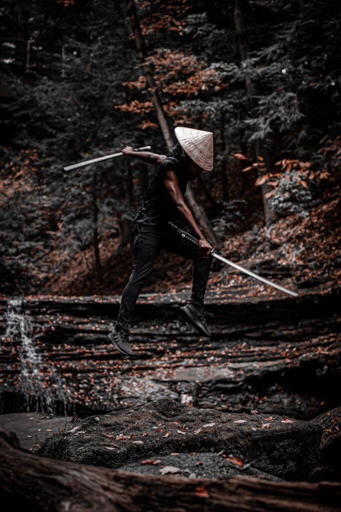 pexels-photo-5971323 A person in a conical hat jumps mid-air with a sword in a forest setting.